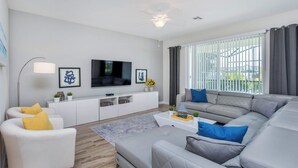 A modern living room with a grey sofa, a white armchair, a flat-screen TV, a ceiling fan, and a large window with blinds.