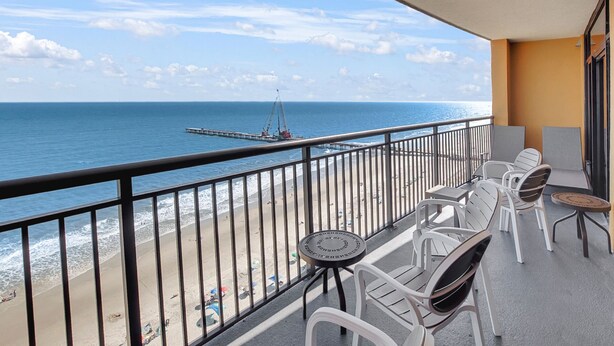 A balcony with white chairs and a small round table, overlooking a beach with a pier and the ocean. - Anderson Ocean Club #1611 (Myrtle Beach)