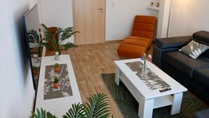A modern living room with a wooden floor, a white coffee table, a black sofa, an orange chair, and a TV on the left. - Vacation apartment König (Arendsee)