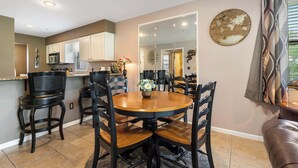 A dining area with a round table and chairs, a kitchen in the background, and a mirror on the wall.