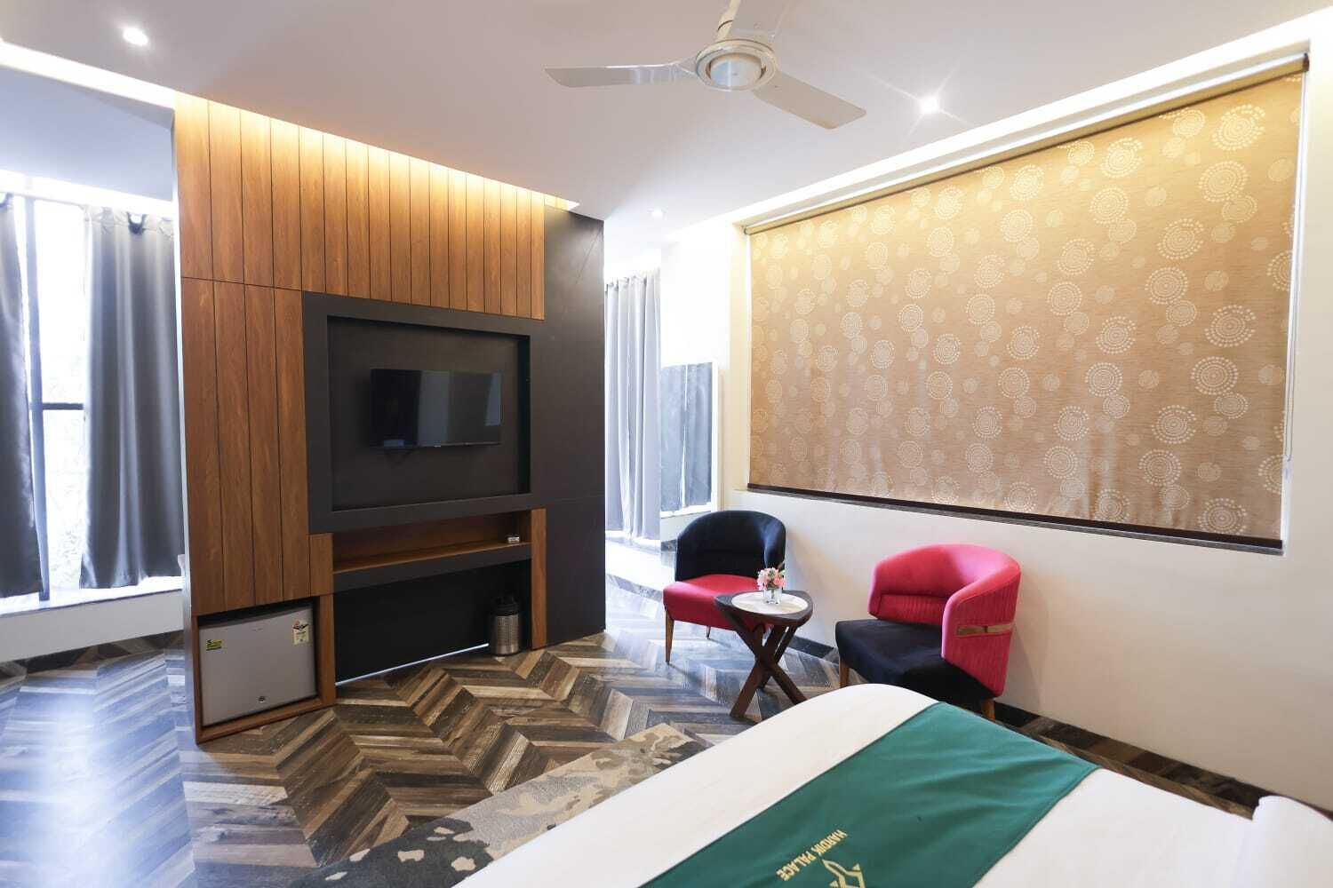 Habitación de hotel con una cama grande, un televisor montado en un panel de madera, una pequeña mesa con sillas y un ventanal con cortinas.