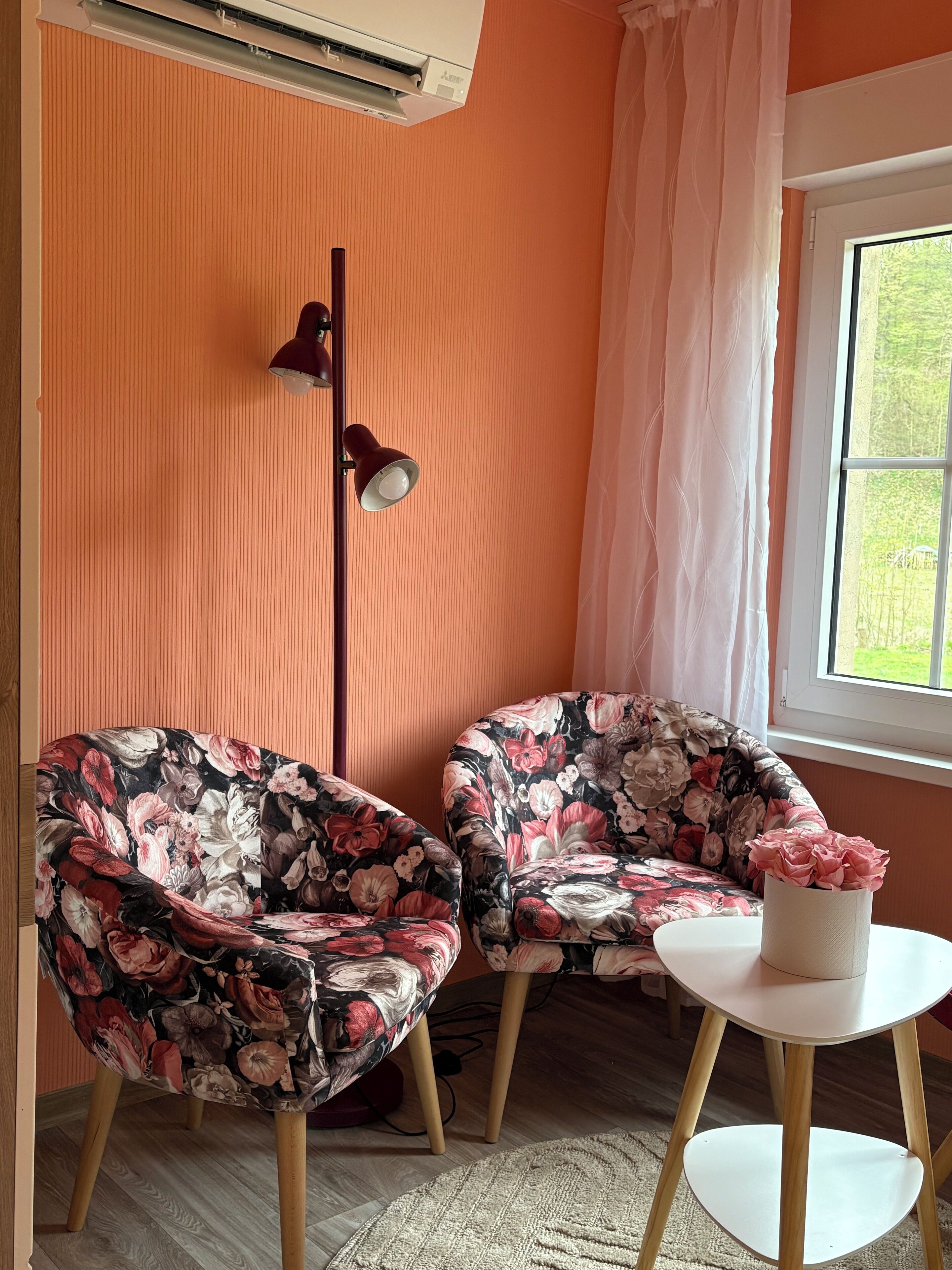 Um quarto com duas cadeiras estampadas com flores, um abajur, uma mesa pequena com um vaso e uma janela com cortinas.