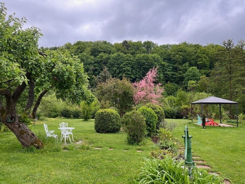 A lush garden with a gazebo, white metal chairs, and a variety of shrubs and trees.