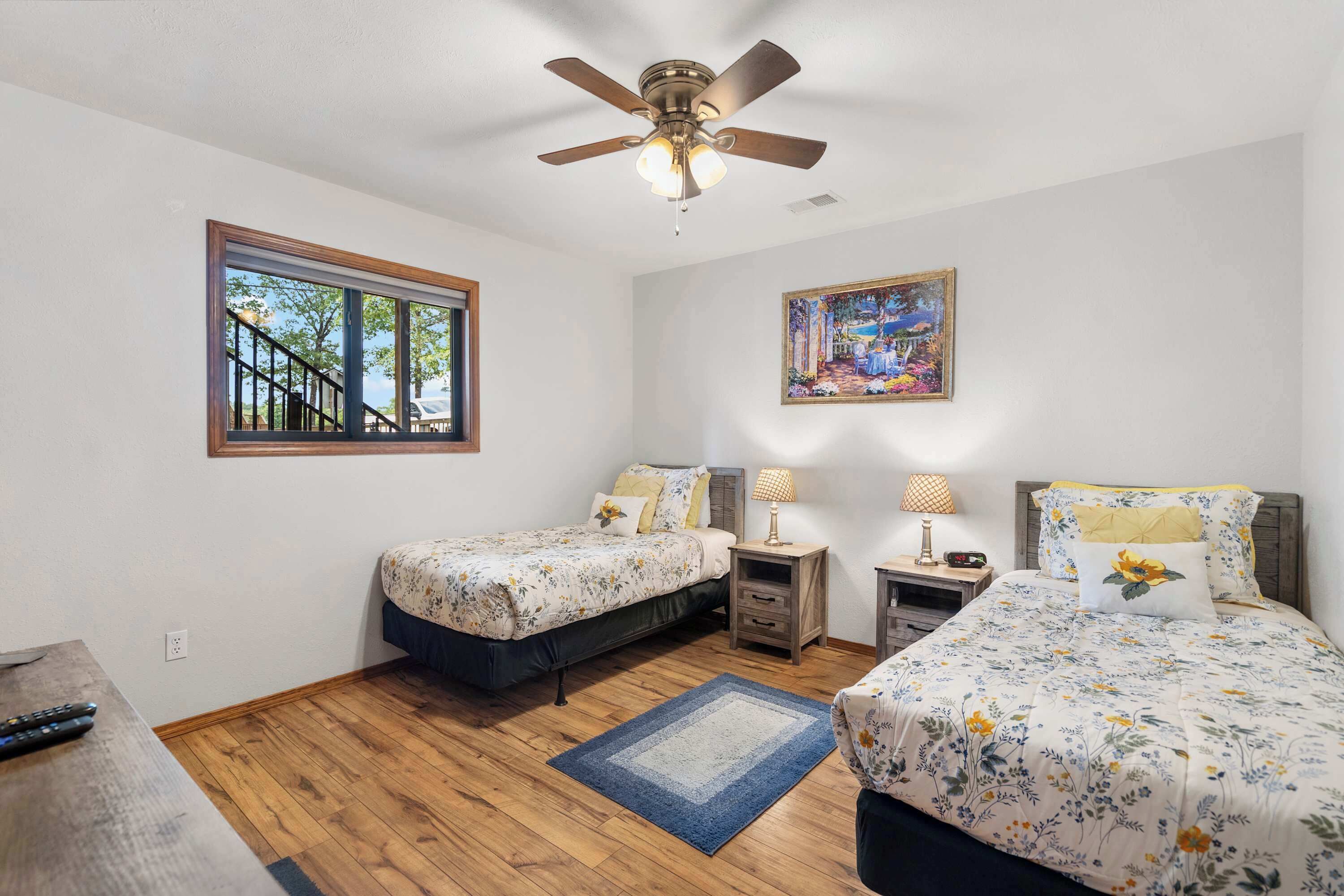 A bedroom with two beds, a ceiling fan, a window with a view, and a painting on the wall.
