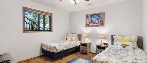 A bedroom with two beds, a ceiling fan, a window with a view, and a painting on the wall.