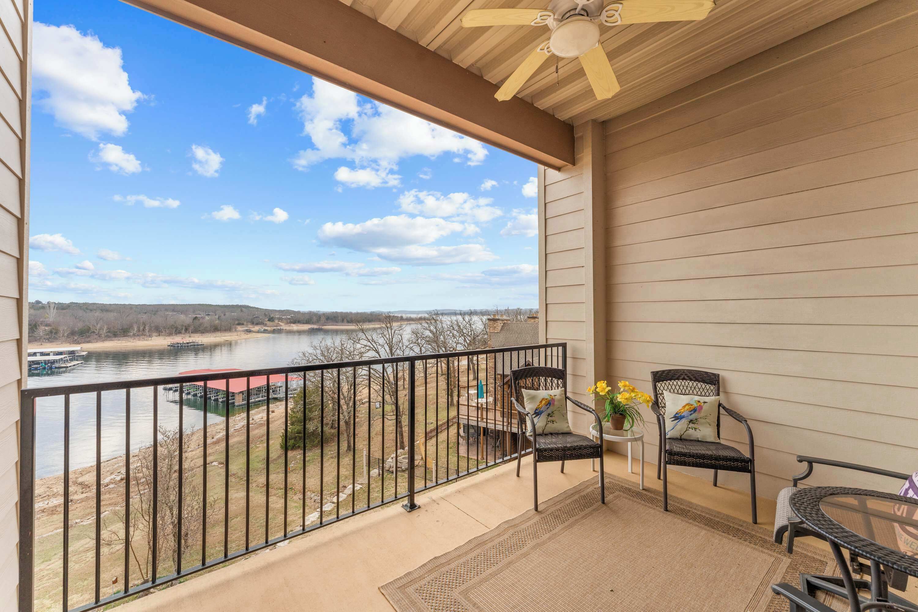 A balcony with a view of a river, two chairs, a table, and a ceiling fan.