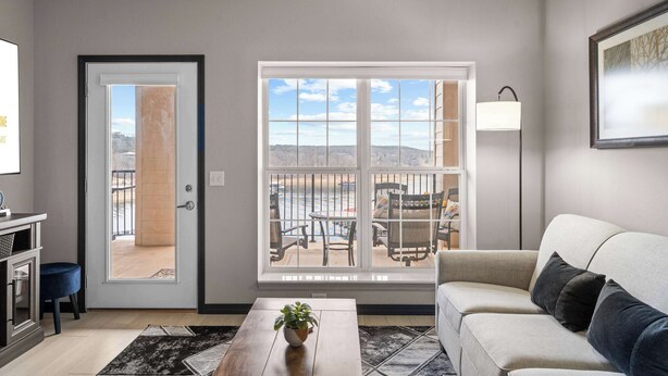 A living room with a sofa, a coffee table with a plant, a window with a view of outdoor furniture, and a door leading outside. - Waters Edge Retreat - For 6! (Branson)
