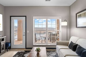 A living room with a sofa, a coffee table with a plant, a window with a view of outdoor furniture, and a door leading outside. - Waters Edge Retreat - For 6! (Branson)