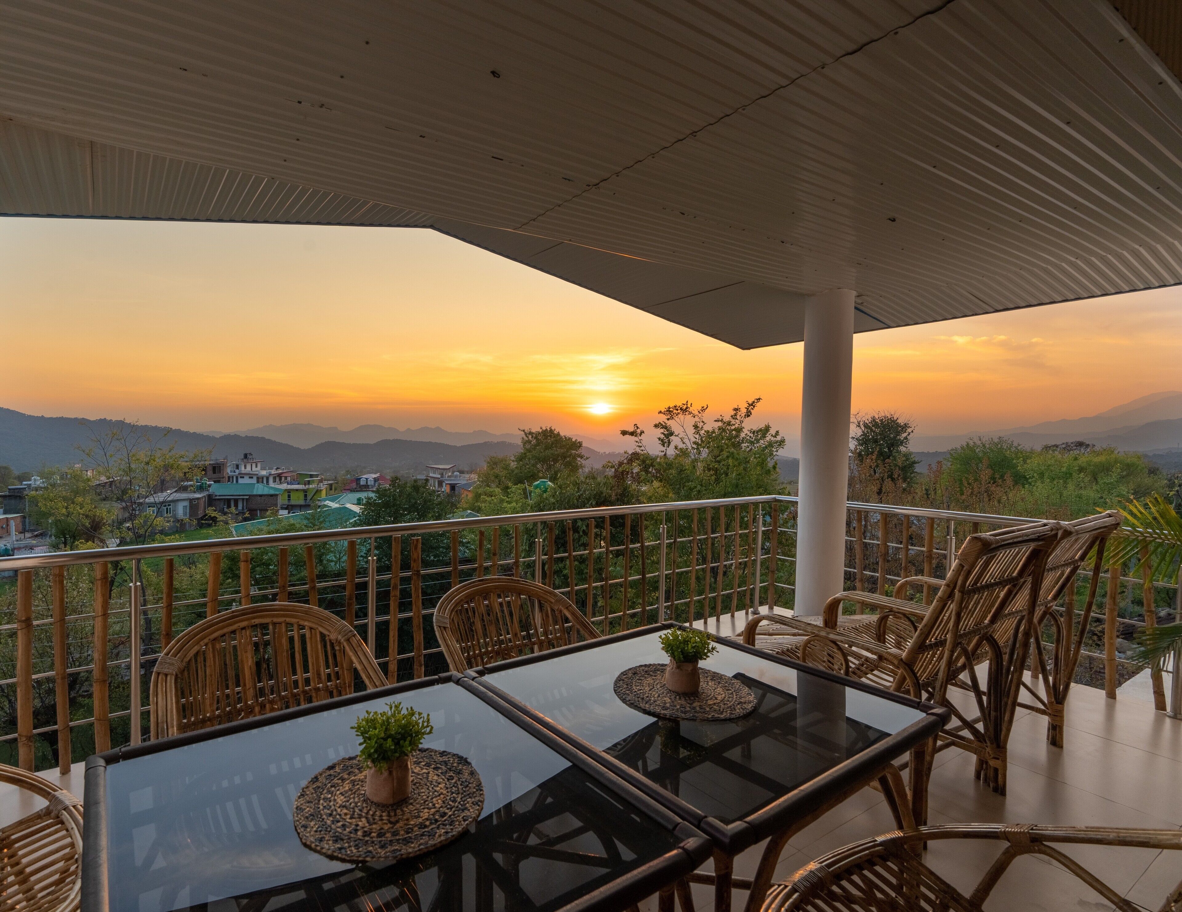 A terrace with a glass table and wicker chairs, overlooking a sunset with mountains in the distance.