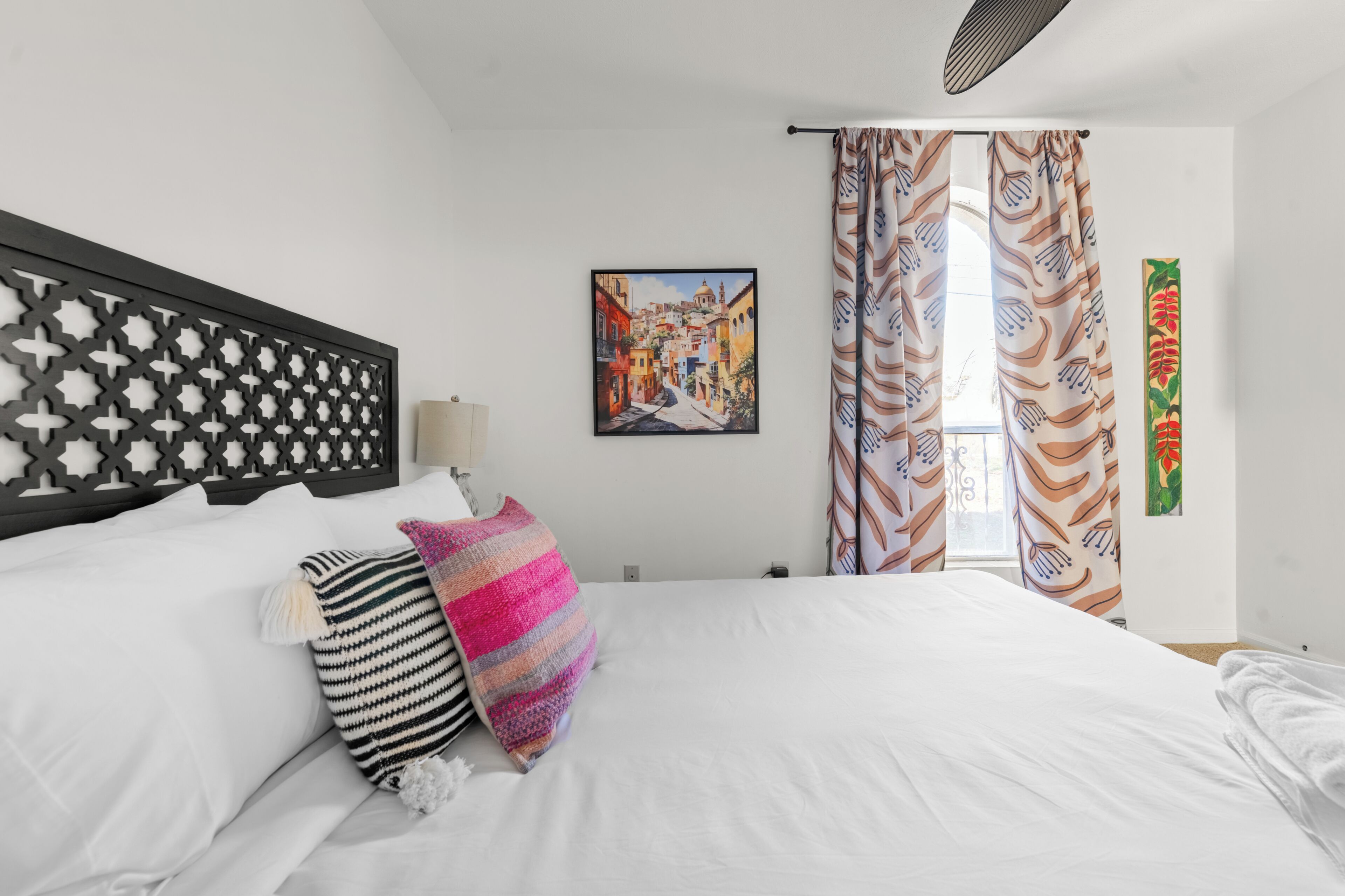 A hotel room with a bed, a decorative headboard, a painting, curtains, and a wall-mounted light.