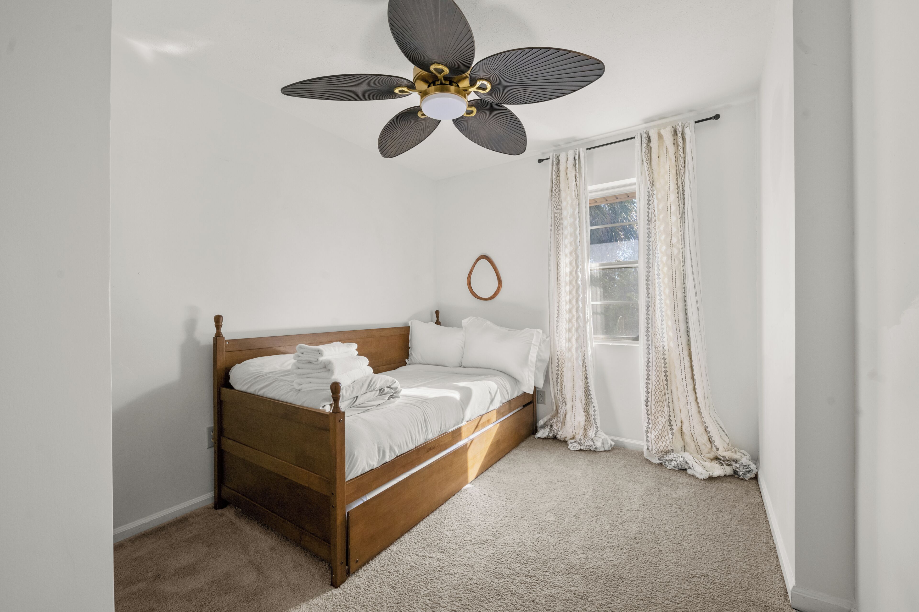 A bedroom with a wooden bed, a ceiling fan, a window with curtains, and a carpeted floor.