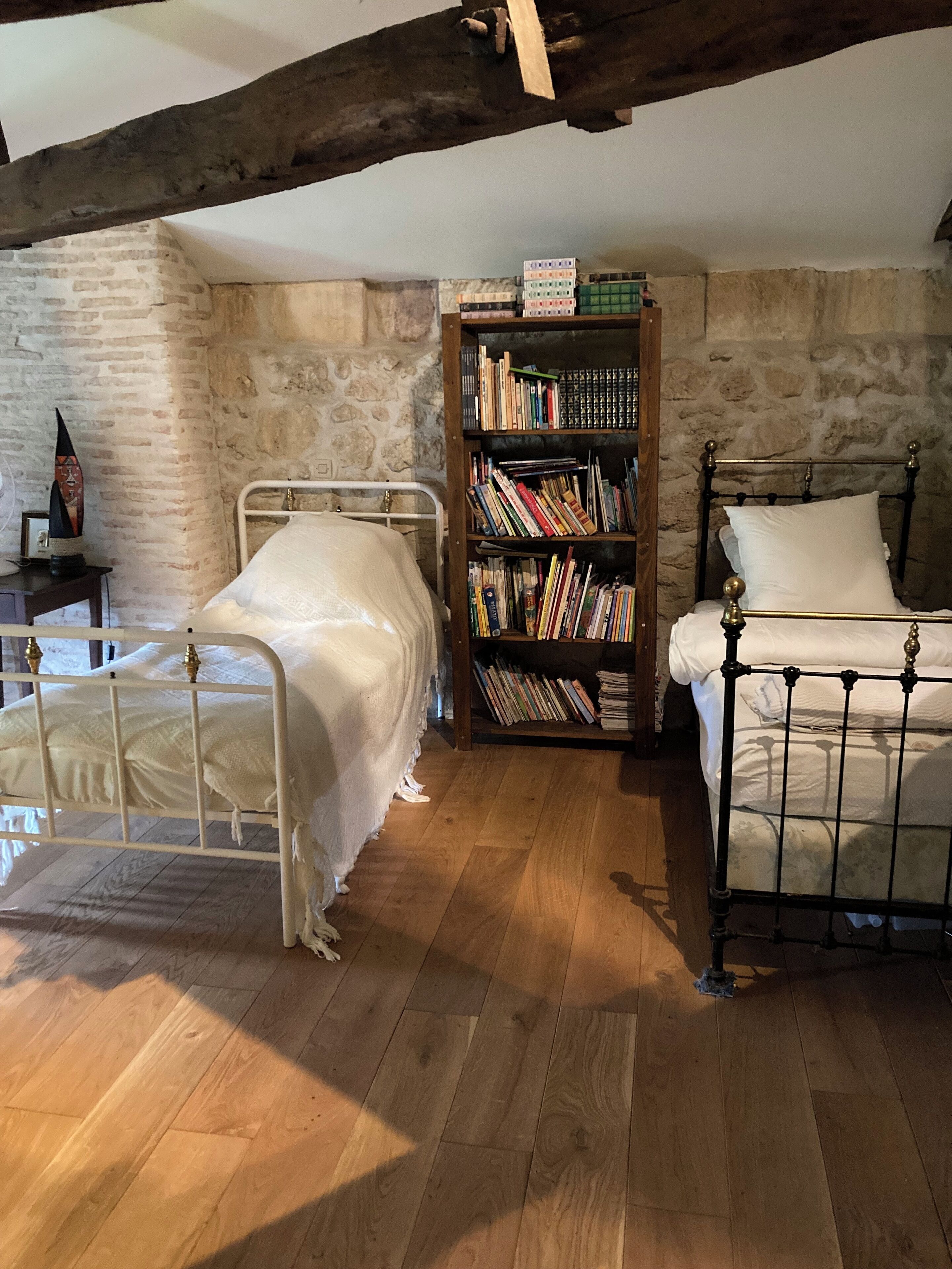 A rustic room with two metal beds, a wooden floor, a stone wall, and a bookshelf filled with books.
