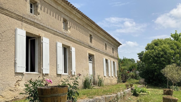 Un bâtiment rustique en pierre, doté de nombreuses fenêtres et d’un jardin agrémenté de plantes et d’un tonneau en bois.