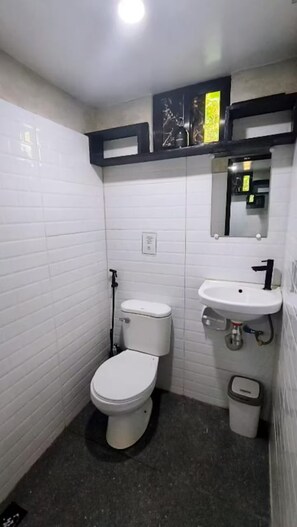A small, compact bathroom with white tiled walls, a white toilet, a white sink, and a trash bin. - El Nido Lofts (El Nido)