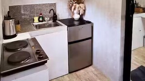 A compact kitchen with a stove, sink, and a small refrigerator. There is a shelf above the sink holding various kitchen items.