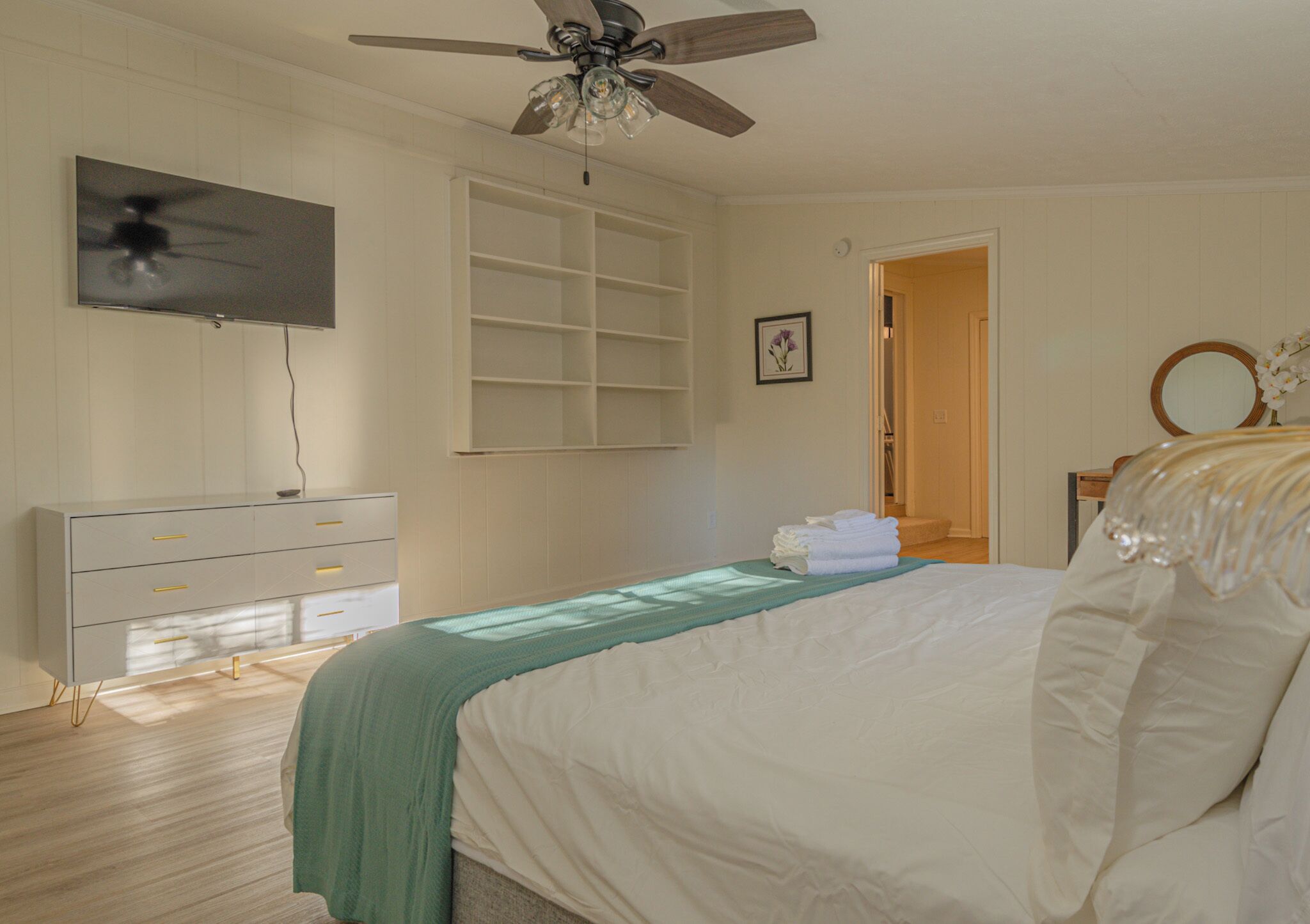 A bedroom with a bed, a ceiling fan, a TV, a dresser, and a door leading to another room.