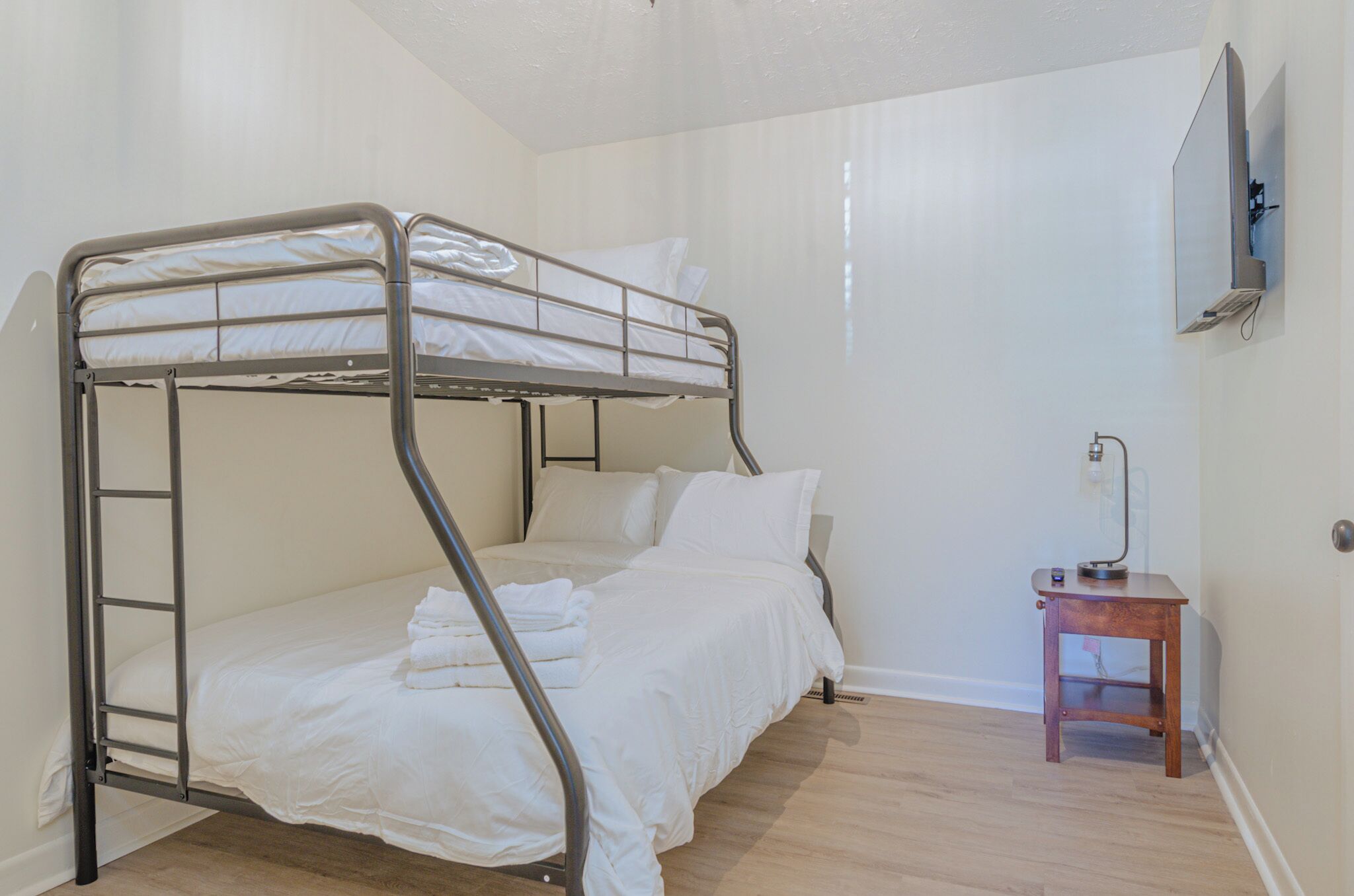 Una cama litera con ropa de cama blanca, una pequeña mesa de madera con una lámpara y un televisor fijado a la pared.
