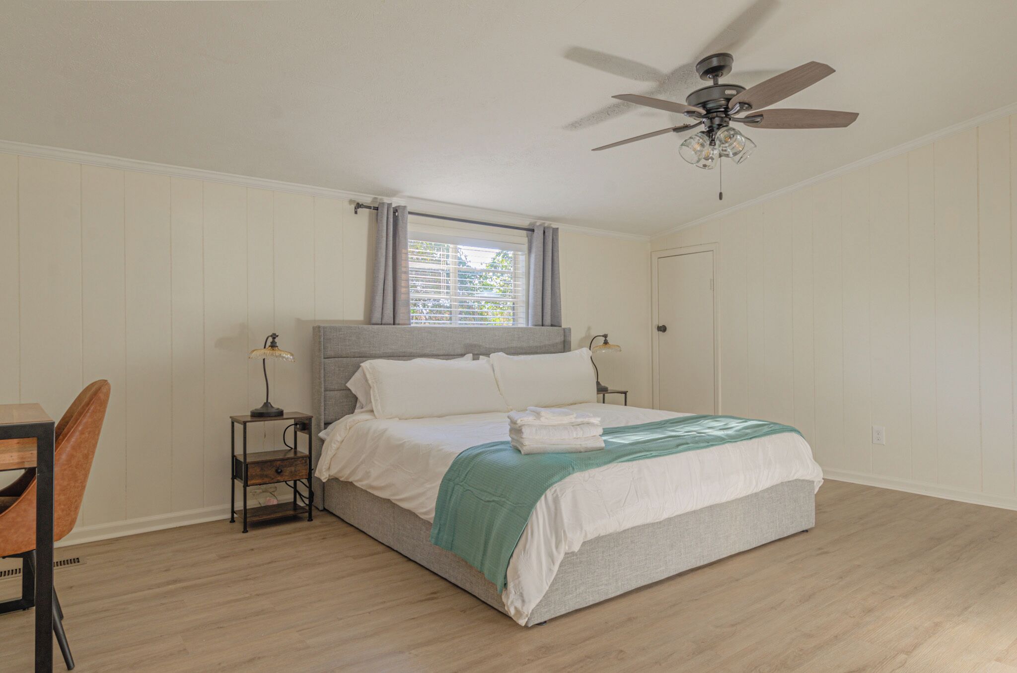A bedroom with a bed, a desk, a chair, a window with curtains, and a ceiling fan.