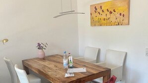 Un coin repas avec une table en bois, des chaises blanches et une chaise rouge. Sur la table, on trouve un vase avec des fleurs, une bouteille et un emballage de jus de fruits.