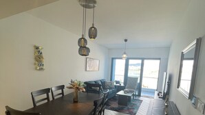 Un salon spacieux avec une grande table à manger, des chaises en bois foncé et un canapé. Des luminaires suspendus sont accrochés au plafond, une grande fenêtre laisse entrer la lumière naturelle et offre une vue sur l’extérieur.