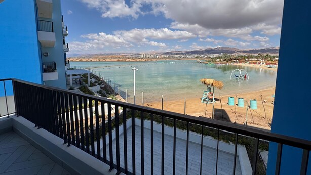 Un balcon donnant sur une plage et la mer, un parasol en chaume et une bouée de sauvetage.