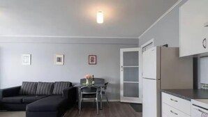 Een moderne woonkamer met een zwarte hoekbank, een eettafel met stoelen en een keuken met witte kasten en een koelkast.