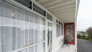 Een overdekt balkon met een tafel en stoelen, grote ramen met gordijnen en een decoratief wandpaneel.