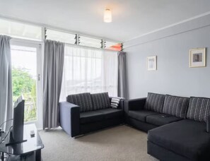 Ein modernes Wohnzimmer mit einem Ecksofa, einem Flachbildfernseher und einem großen Fenster mit Vorhängen.