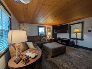 Un salon chaleureux avec un plafond en bois, un canapé, une table basse sur laquelle est posé un jeu d’échecs, un téléviseur et une lampe.