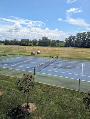 Una cancha de tenis exterior con red, rodeada por una valla y árboles.