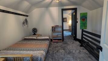 Un dormitorio con una cama, una mesita de noche, una silla, un ventilador, un estante con libros, un cuadro y una lámpara en el techo.