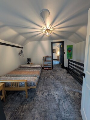 A bedroom with a bed, a bedside table, a chair, a fan, a shelf with books, a painting, and a ceiling light fixture.