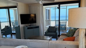 A modern living room with a white sofa, a dining table, and a TV. There is a balcony with a view of the ocean.