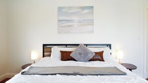 Una cama bien hecha con ropa blanca, un plaid gris y dos almohadas marrones. En la pared sobre la cama hay un cuadro con una escena de playa.