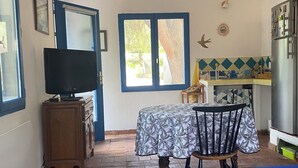 A compact room with a bed, a television, a dining table with chairs, a kitchen area, and a window with a view of greenery.