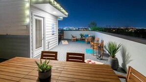 A rooftop patio with a wooden table and chairs, potted plants, and a view of the city skyline at night. - Nashville Skyline View with a Rooftop Deck that Sleeps 12 People (Nashville)