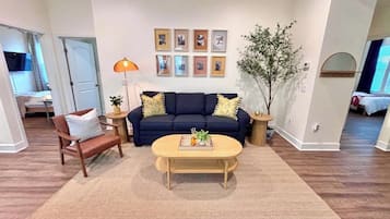 A living room with a sofa, coffee table, and a rug. There are framed pictures on the wall and a plant.