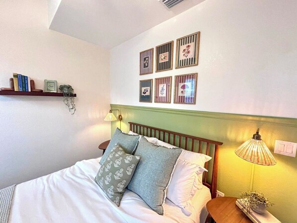A neatly made bed with pillows, a bedside lamp, and a wall-mounted shelf with books and a small plant.