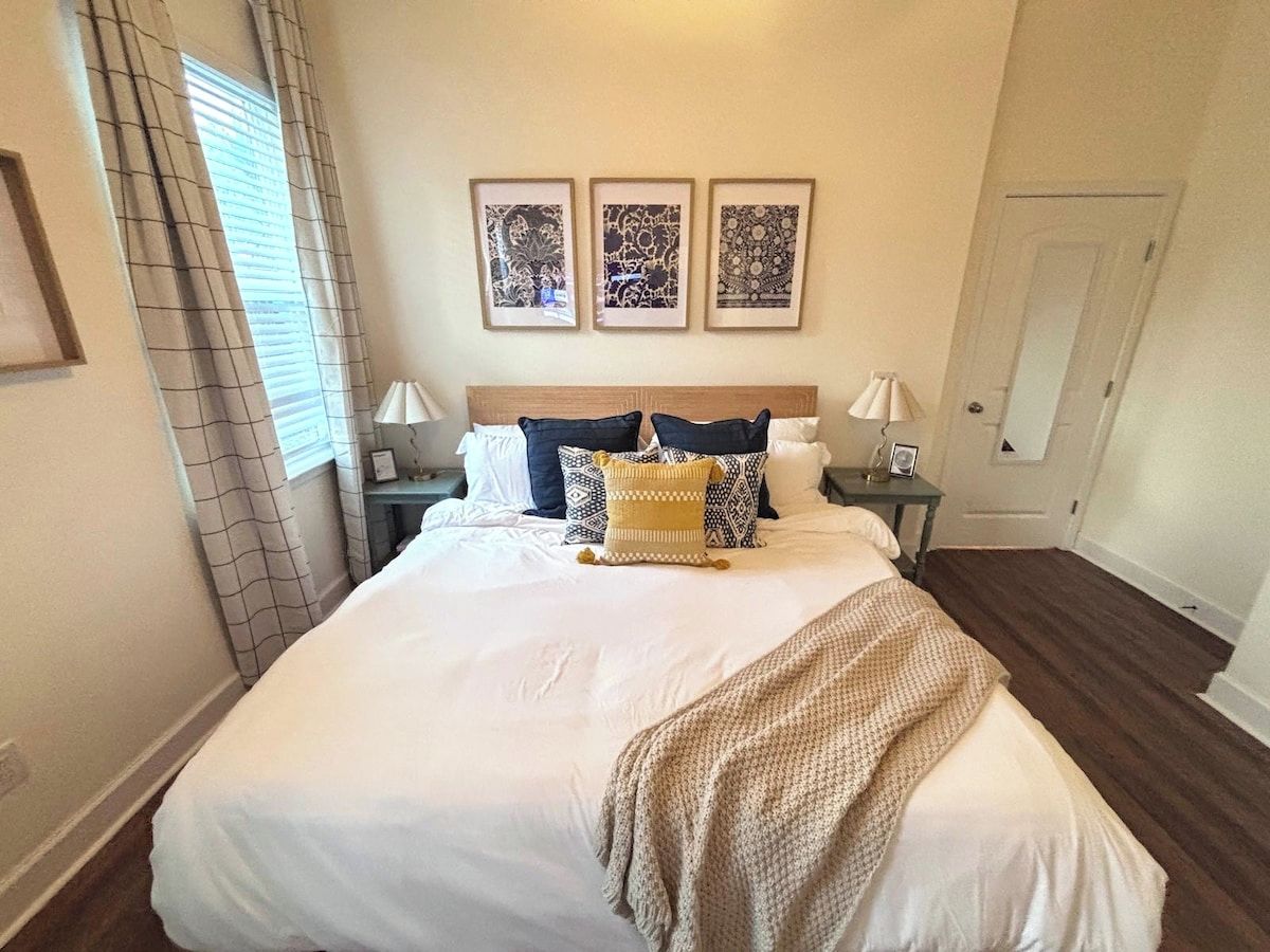 A neatly made bed with a white comforter and a beige throw blanket, flanked by two bedside tables with lamps, a window with curtains, and three framed wall art pieces above the bed.