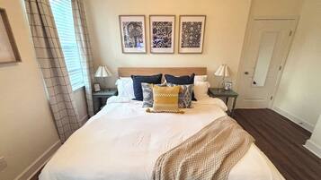 A neatly made bed with a white comforter and a beige throw blanket, flanked by two bedside tables with lamps, a window with curtains, and three framed wall art pieces above the bed.