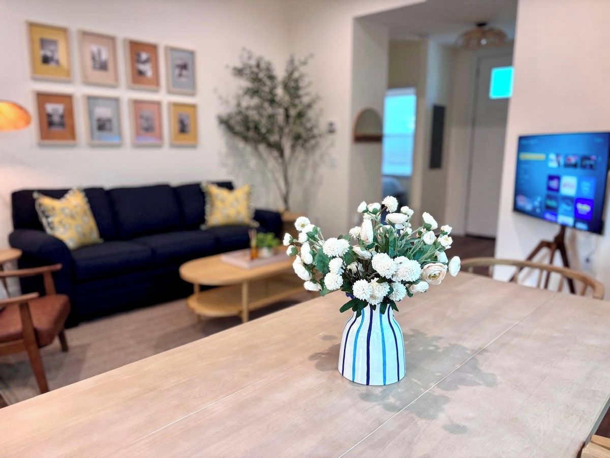Una mesa de comedor con un florero, un sofá, una mesa de centro y un televisor al fondo.