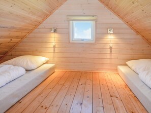 Une pièce aux murs en bois et au plafond en pente, avec deux lits dotés de draps blancs, une petite fenêtre et des lampes fixées au mur.