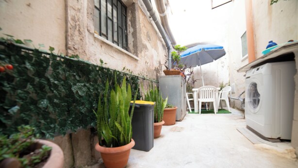 A narrow outdoor corridor with potted plants, a washing machine, and a chair under a blue umbrella. - Cozy vacation home, modern amenities and guaranteed relaxation. We are waiting for you! (Siracusa)