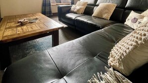 A living room with a black leather sofa, a wooden coffee table, and a window with blue curtains. - Rustic barn Cottage in a rural village with traditional open fireplace sleeps 5. (Cockermouth)