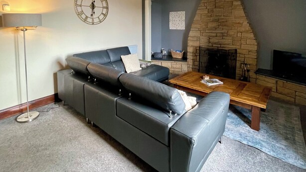 A living room with a stone fireplace, a black leather sofa, a wooden coffee table, and a clock on the wall. - Rustic barn Cottage in a rural village with traditional open fireplace sleeps 5. (Cockermouth)