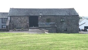A stone building with a dark entrance, a small porch, and a grassy lawn in front. - Rustic barn Cottage in a rural village with traditional open fireplace sleeps 5. (Cockermouth)