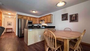 A compact kitchen with wooden cabinets, a stainless steel refrigerator, and a dining table with chairs. - Ski-In Stone Creek Condo - Hot Tub - 3 bedroom - Pet Friendly (Fernie)