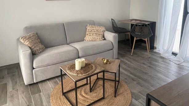 A living room with a grey sofa, two side tables, a coffee table, and a dining area with a chair and table. - Studio near La Rochelle (Dompierre-sur-Mer)