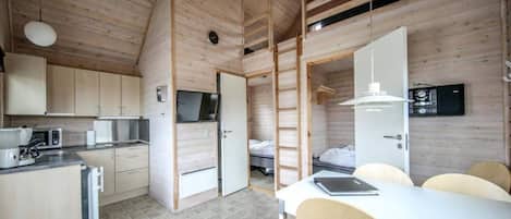 Cozinha compacta e moderna, com armários de madeira, área de jantar com mesa e cadeiras brancas, e um mezanino com quarto acessado por escada.
