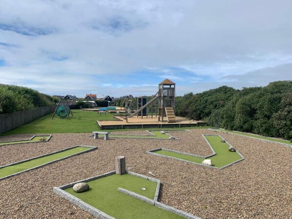 Un parque infantil con una estructura de escalada y un tobogán, rodeado de un área ajardinada con senderos de grava y zonas verdes.